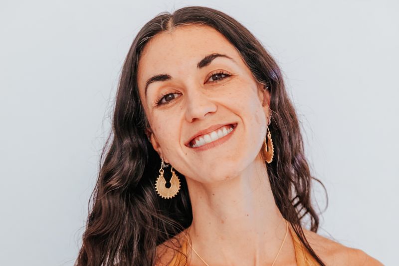 Una mujer sonriente con cabello largo y oscuro y aretes de oro, fotografiada contra un fondo claro.