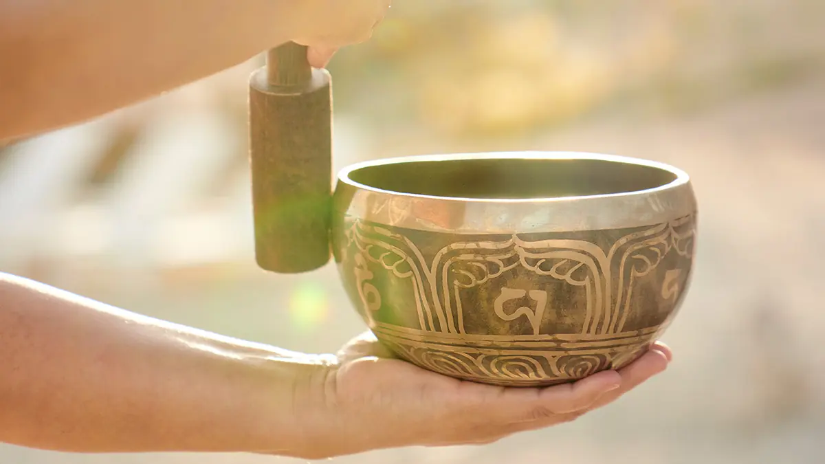Hands holding ornate Tibetan singing bowl with wooden mallet, glowing softly in warm sunlight.