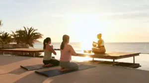 Two women practice yoga at sunset while instructor uses singing bowl beside oceanfront infinity pool.