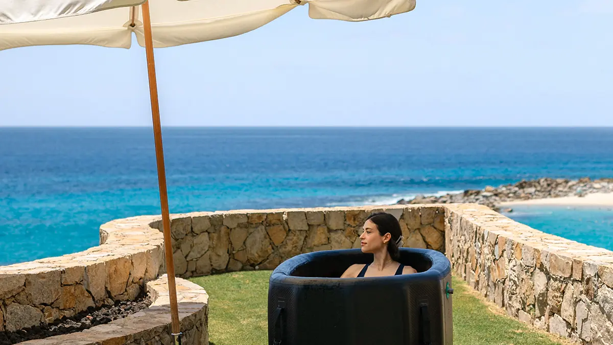 Person relaxes in a black soaking tub outdoors, under umbrella, overlooking blue ocean view.