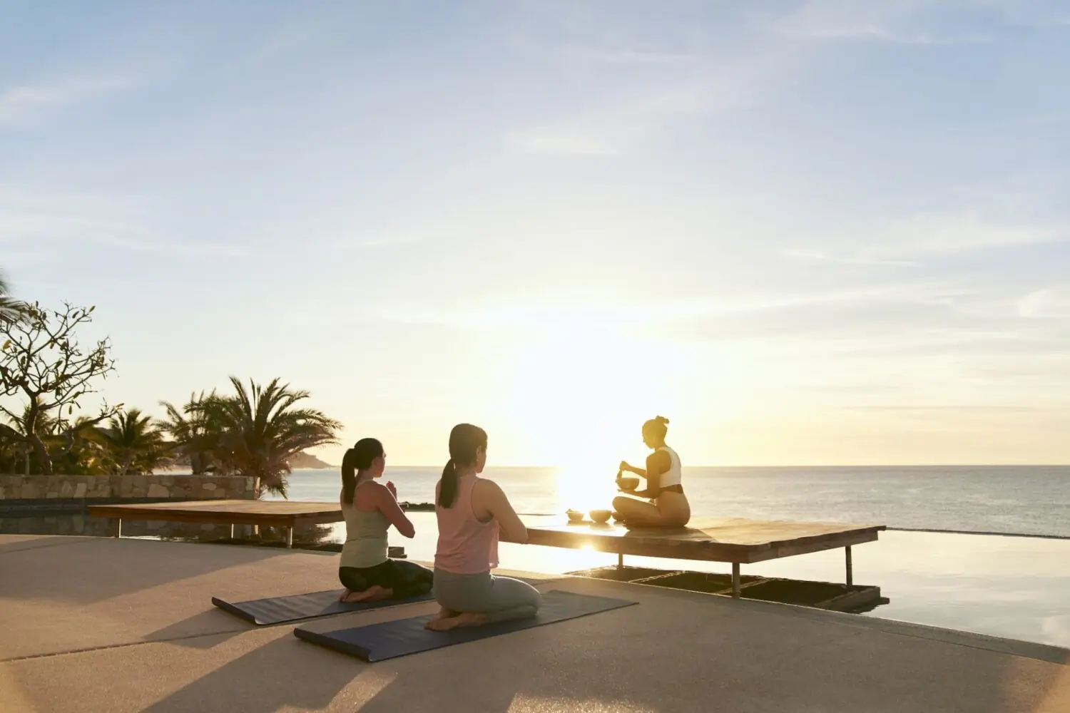 Three people practice sunrise yoga on mats by infinity pool, ocean horizon, and palm trees in background.