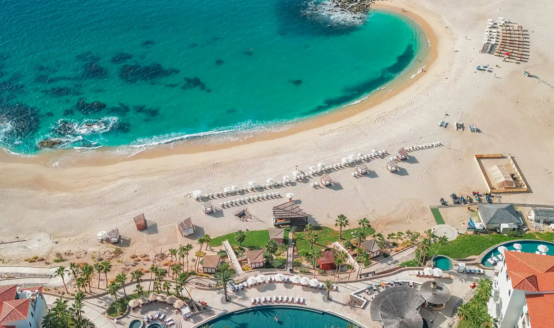 Vista aérea de un complejo tropical con aguas turquesas, sombrillas de playa blancas, piscinas y edificios con techos rojos.