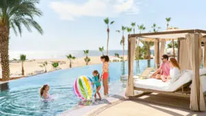 Niños jugando con una gran pelota de playa de colores en la piscina, padres relajándose en una cabaña junto a las palmeras.