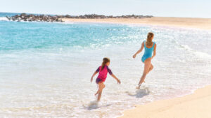 Woman and child joyfully run along sandy beach shoreline, splashing in ocean waves under sunshine.