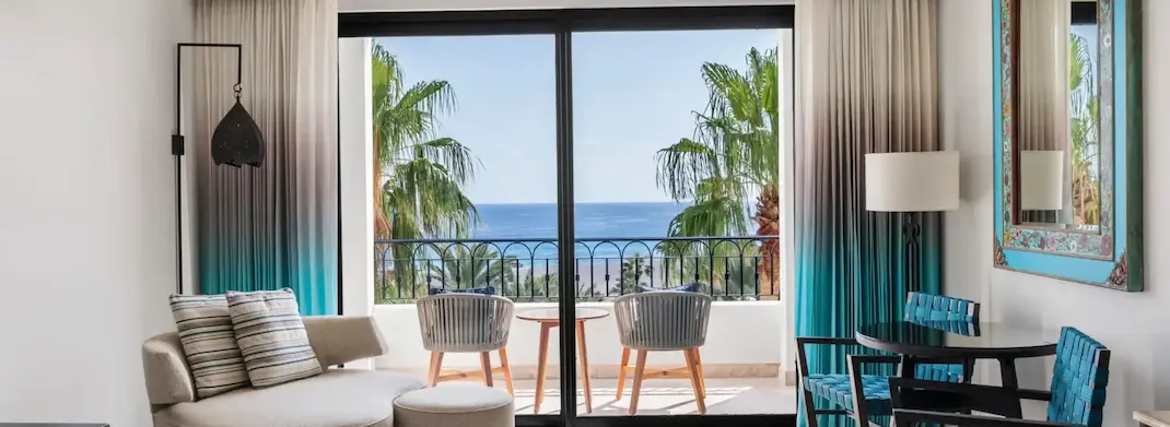 Modern hotel suite with seating area and balcony overlooking palm trees and the ocean.
