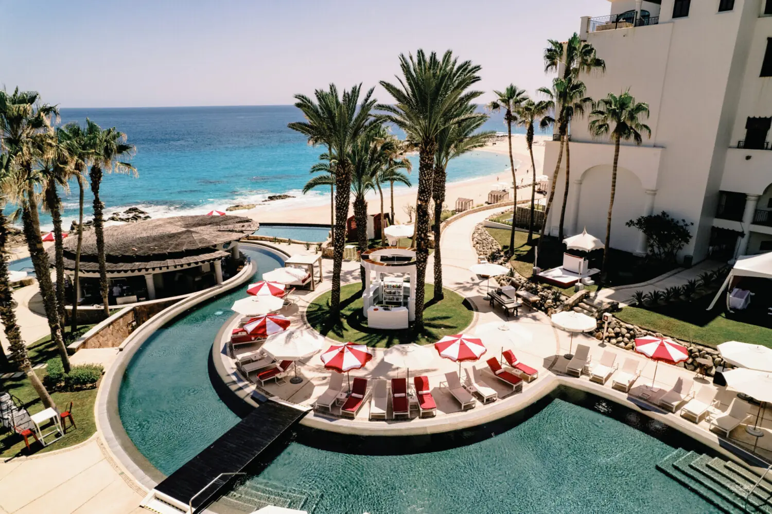 Resort swimming pool area with loungers, umbrellas, palm trees, and ocean beach view under sunny sky.