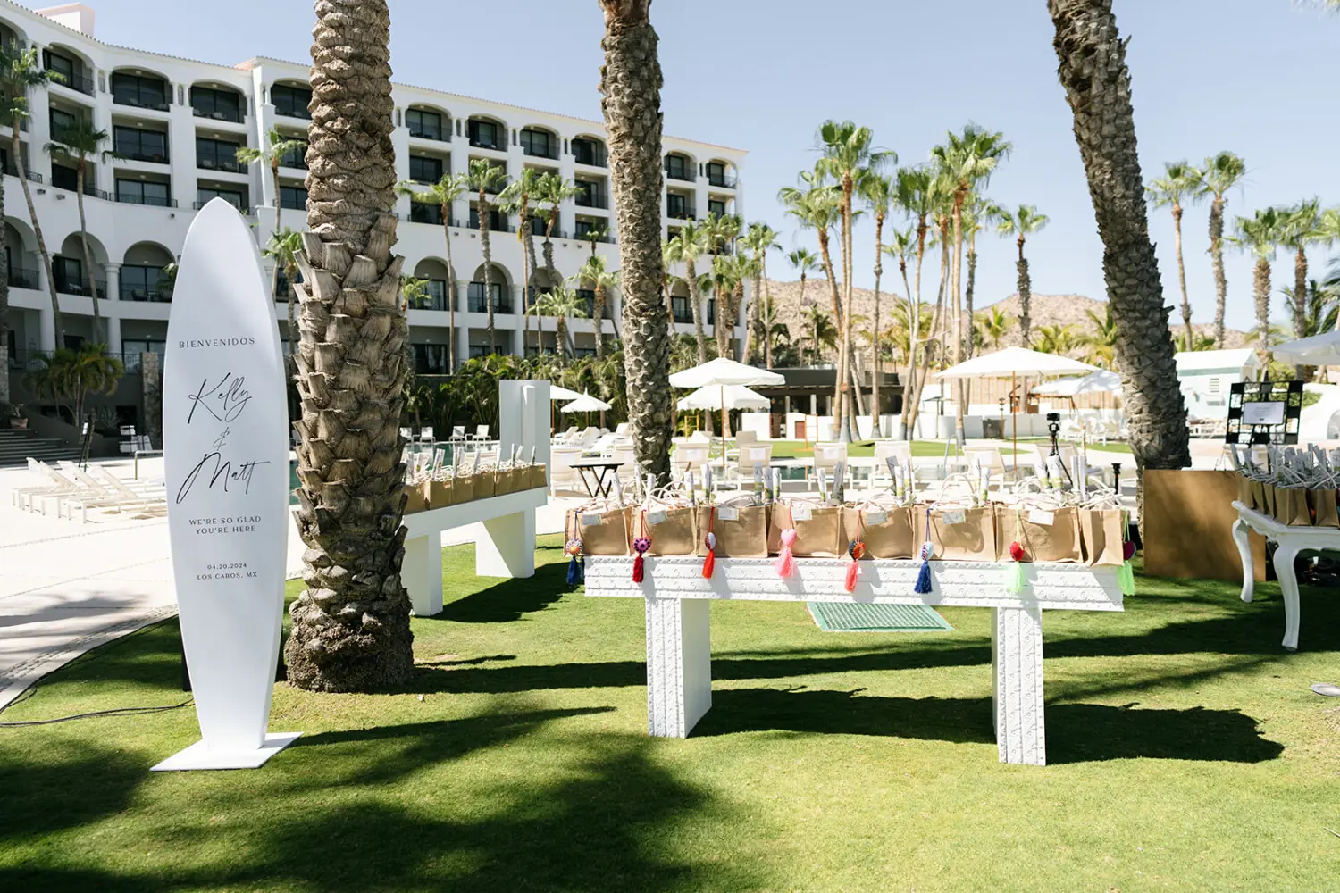 Outdoor wedding welcome setup with personalized sign, gift bags on tables, palm trees, and resort backdrop.
