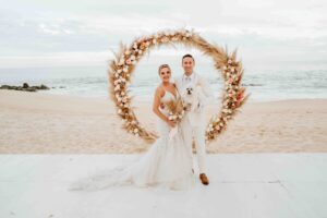 Bride and groom stand on beach in wedding attire under circular floral decoration facing camera.