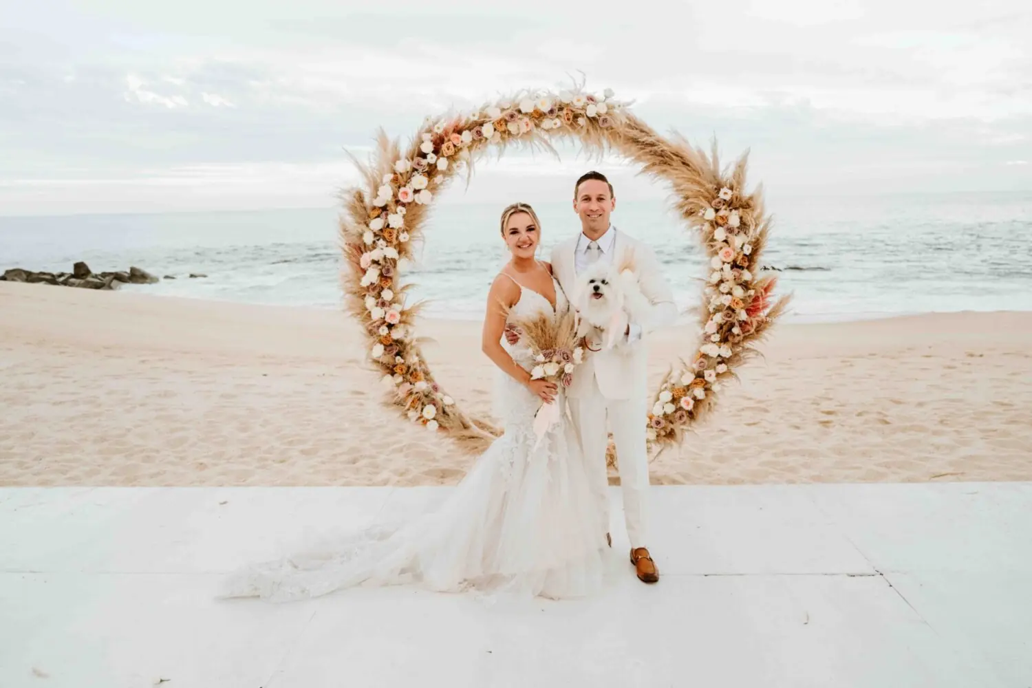 La novia y el novio están de pie en la playa con atuendo de boda bajo una decoración floral circular frente a la cámara.