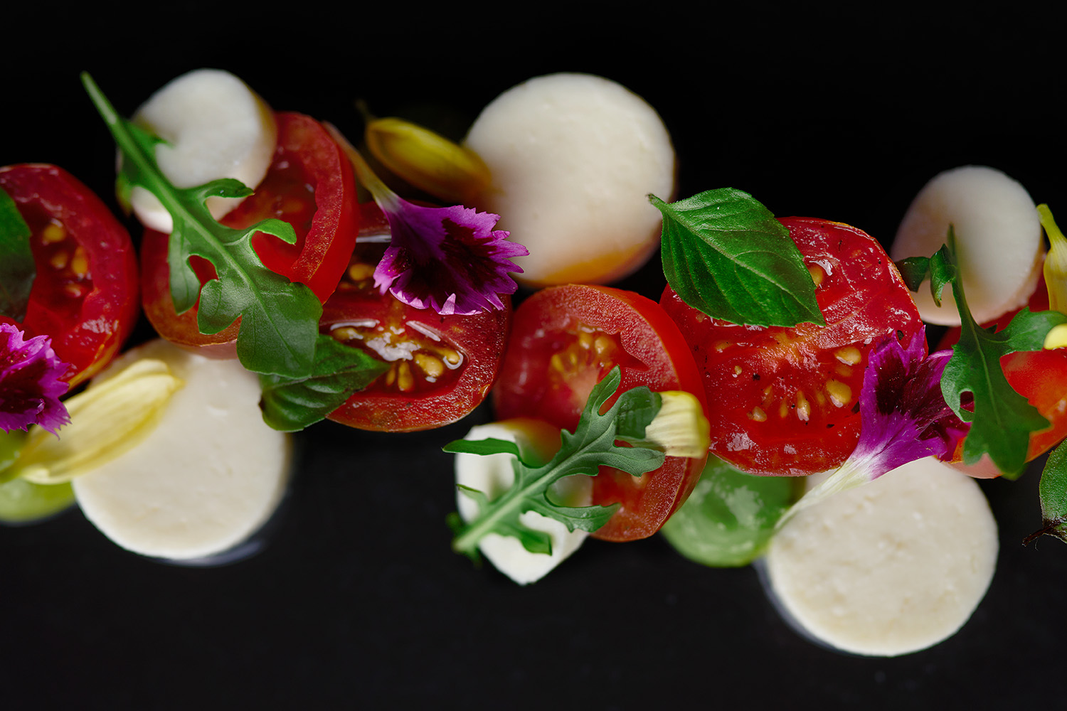 Primer plano de una ensalada estilo Caprese con mozzarella, tomates cherry, hierbas frescas y flores comestibles sobre un fondo oscuro.