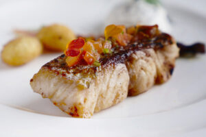 Filete de pescado a la parrilla cubierto con guarnición de tomate cortado en cubitos, servido en plato blanco con patatas y salsa.