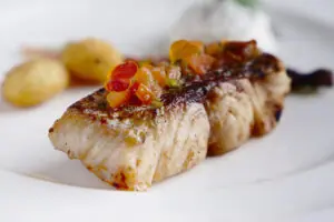Grilled fish fillet topped with diced tomato garnish, served on white plate with potatoes and sauce.