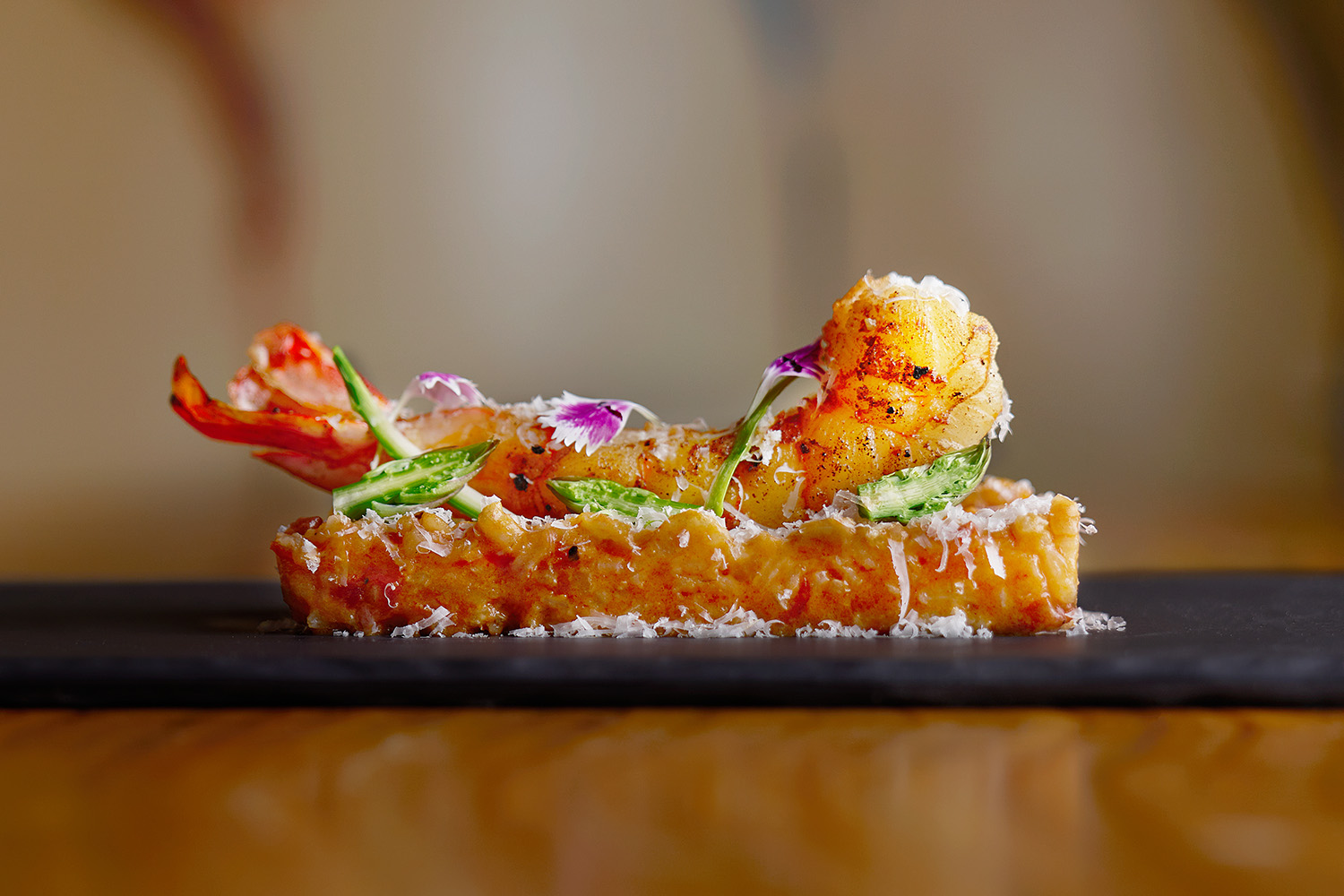 Plato de langosta gourmet elegantemente presentado con hierbas, flores comestibles y queso rallado sobre una superficie oscura.