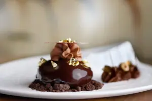Elegant chocolate dessert topped with gold leaf, cream swirl, and nuts on a white plate.