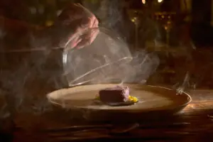 Chef lifts glass dome, releasing aromatic smoke over elegantly plated gourmet dish on rustic restaurant table.