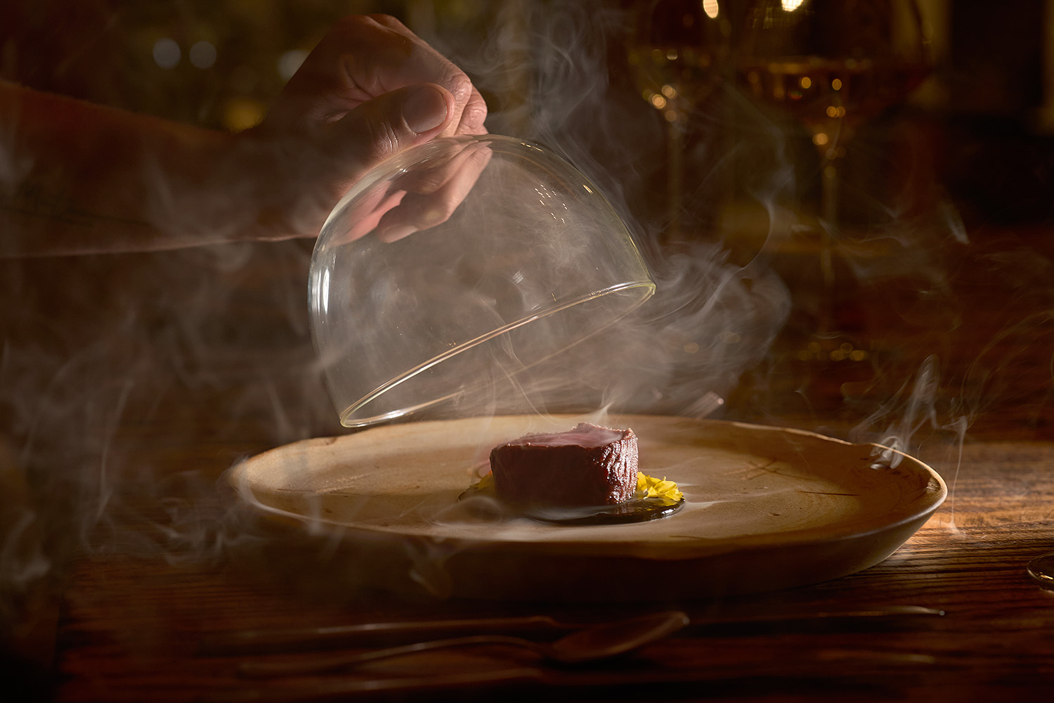 El chef levanta la cúpula de cristal, liberando humo aromático sobre un plato gourmet elegantemente presentado en una mesa de restaurante rústico.