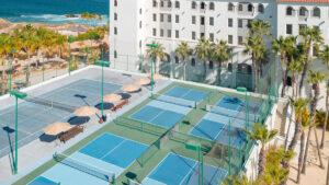Canchas de tenis y pickleball en un complejo turístico frente al mar rodeado de palmeras con un edificio de hotel blanco cerca.