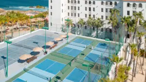 Oceanfront resort tennis and pickleball courts surrounded by palm trees with a white hotel building nearby.