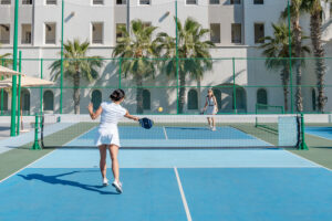 Two women playing pickleball on an outdoor court with palm trees and building background.