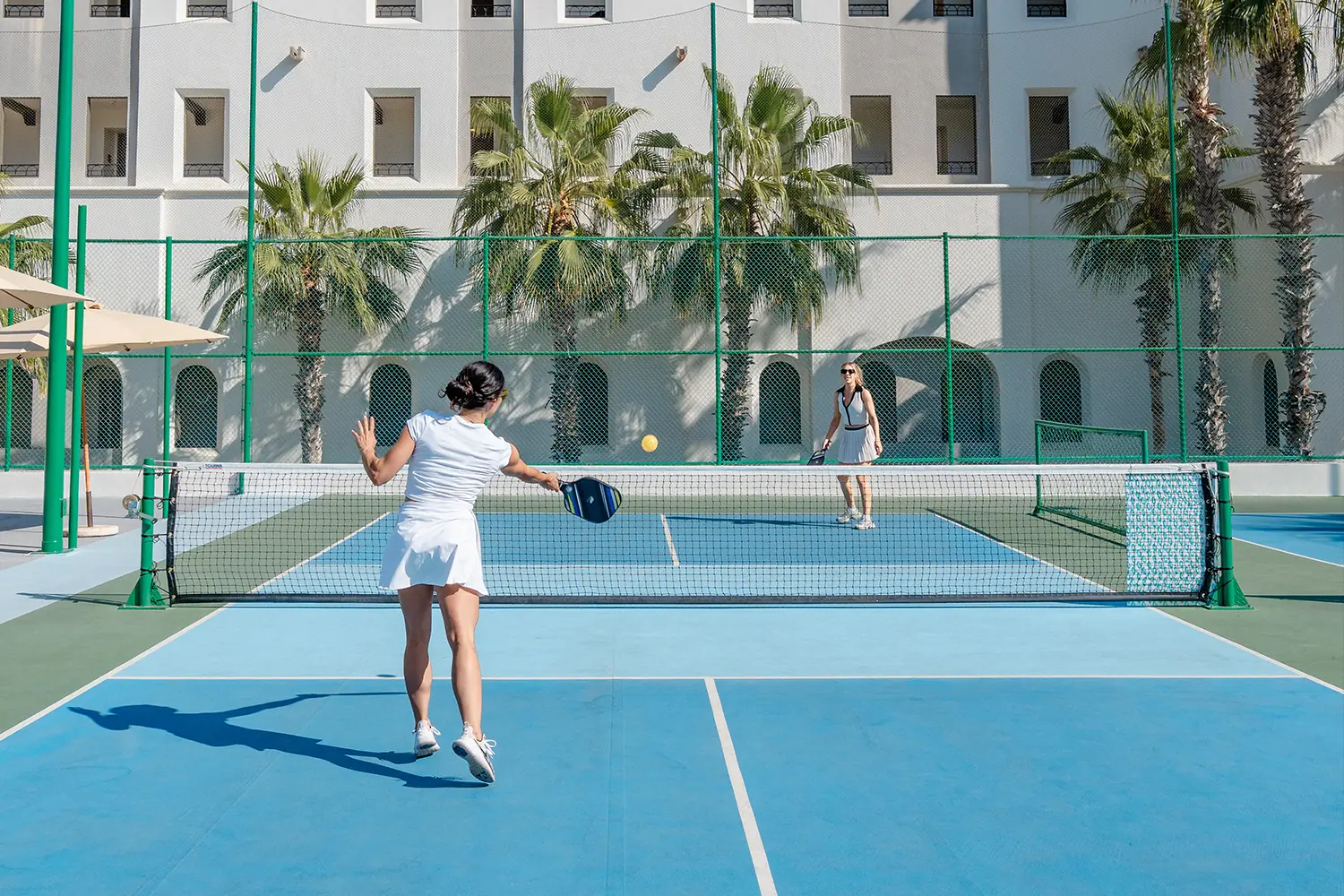 Two women playing pickleball on an outdoor court with palm trees and building background.