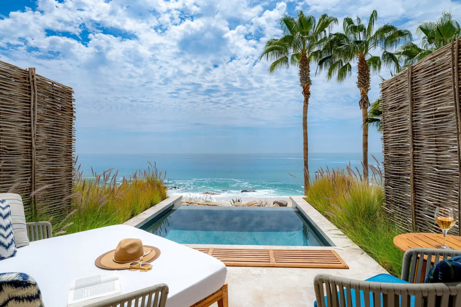 Un patio frente al mar con piscina privada, asientos tipo lounge, palmeras y una vista despejada del mar.