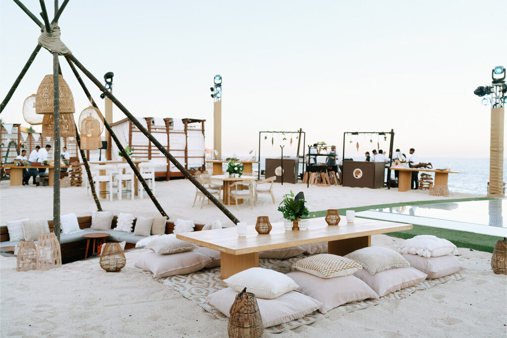 Beachfront event setup with low wooden tables, floor cushions, lanterns, and an ocean view.