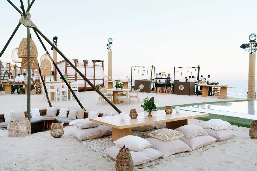 Beachfront event setup with low wooden tables, floor cushions, lanterns, and an ocean view.