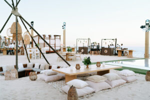 Montaje para eventos frente a la playa con mesas bajas de madera, cojines de piso, linternas y vista al mar.