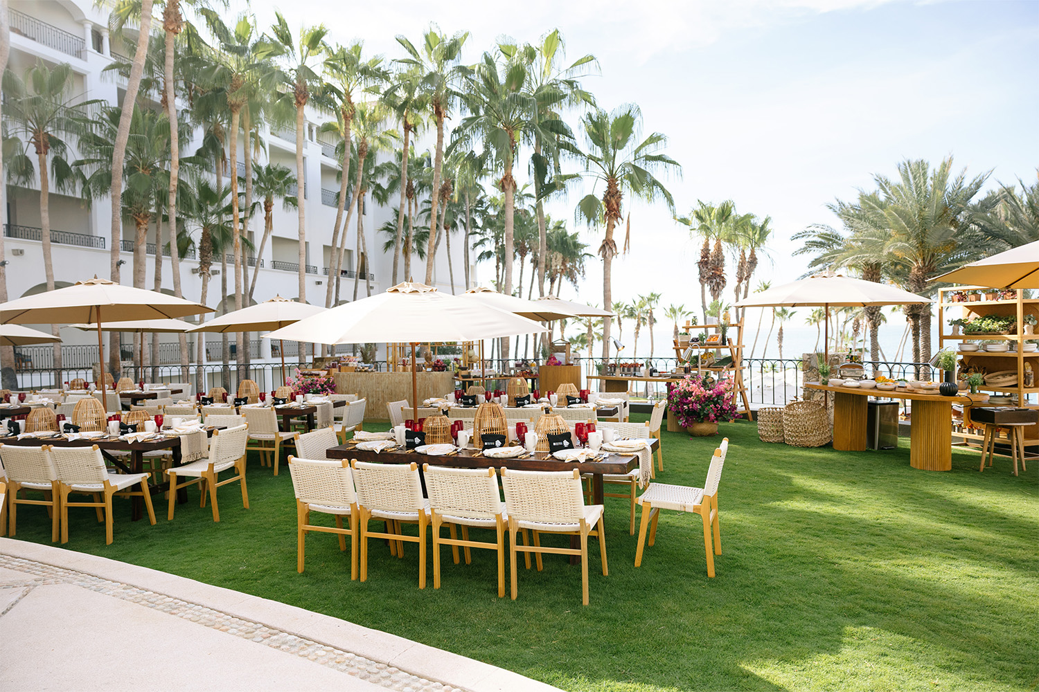 Comedor al aire libre frente a la playa con mesas largas, sombrillas, palmeras y vistas al océano en un resort.