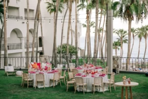 Outdoor garden event with round tables, pink flowers and napkins, string lights, palm trees, and ocean view in background.