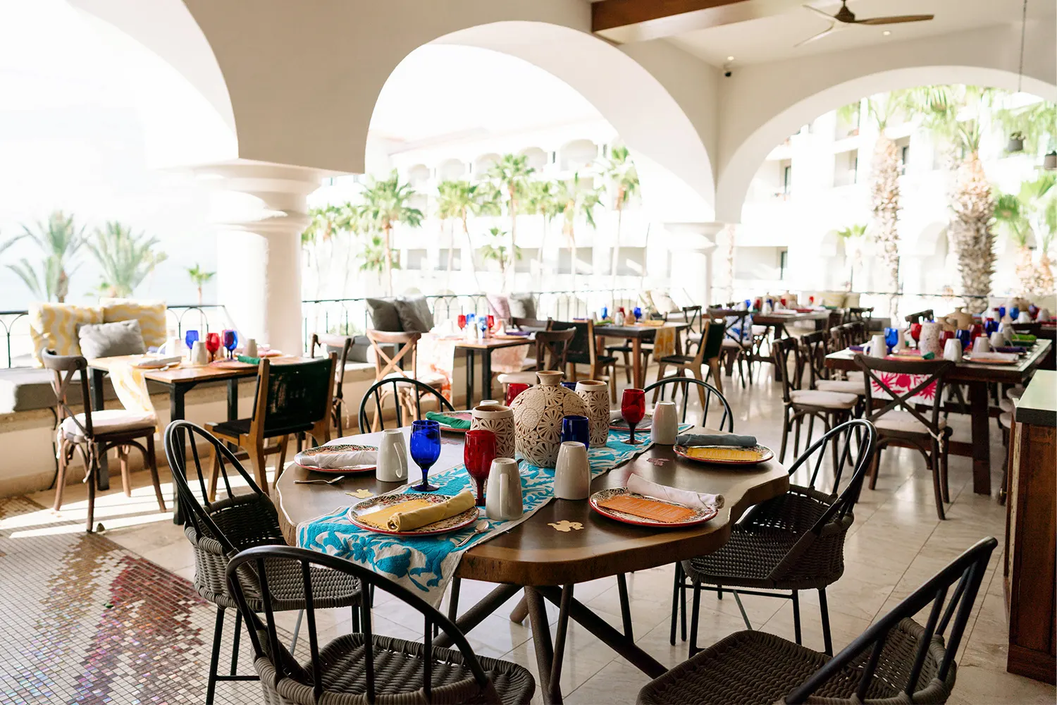 Outdoor dining area with colorful table settings, wicker chairs, and arched roof overlooking palm trees.