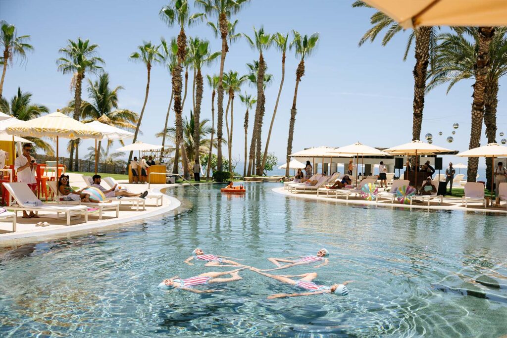 Guests relax by a resort pool with palm trees and loungers as swimmers float in formation under a sunny sky.
