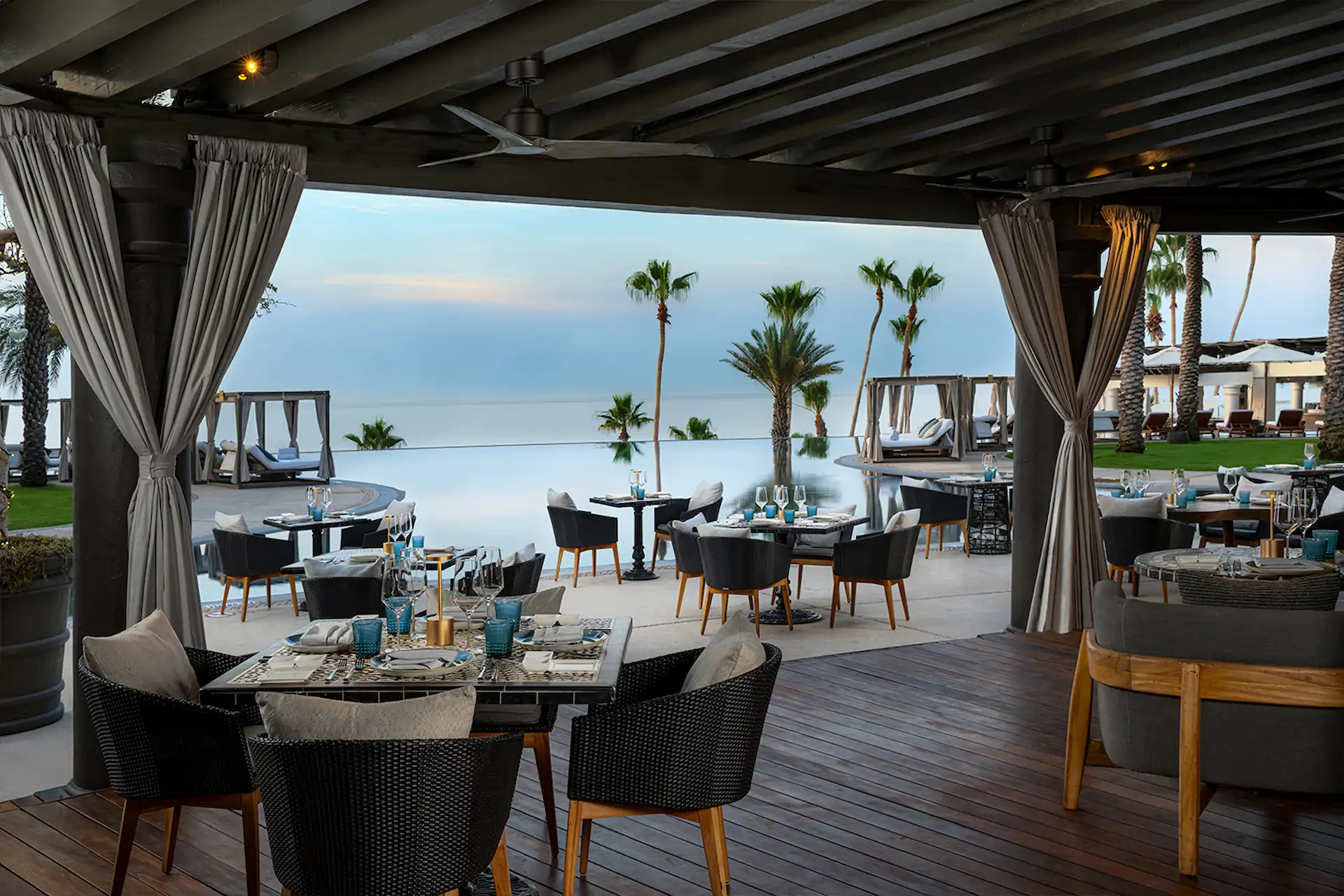 Upscale outdoor dining area at a luxury resort, pergola-covered tables, infinity pool, palm trees & ocean views at sunset.