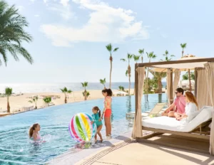Children play with beach ball in infinity pool while adults relax on shaded cabana, palm trees nearby.