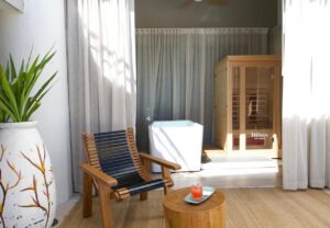 Sala de spa tranquila con silla de madera, mesa pequeña, bañera independiente y sauna privada detrás de cortinas transparentes.