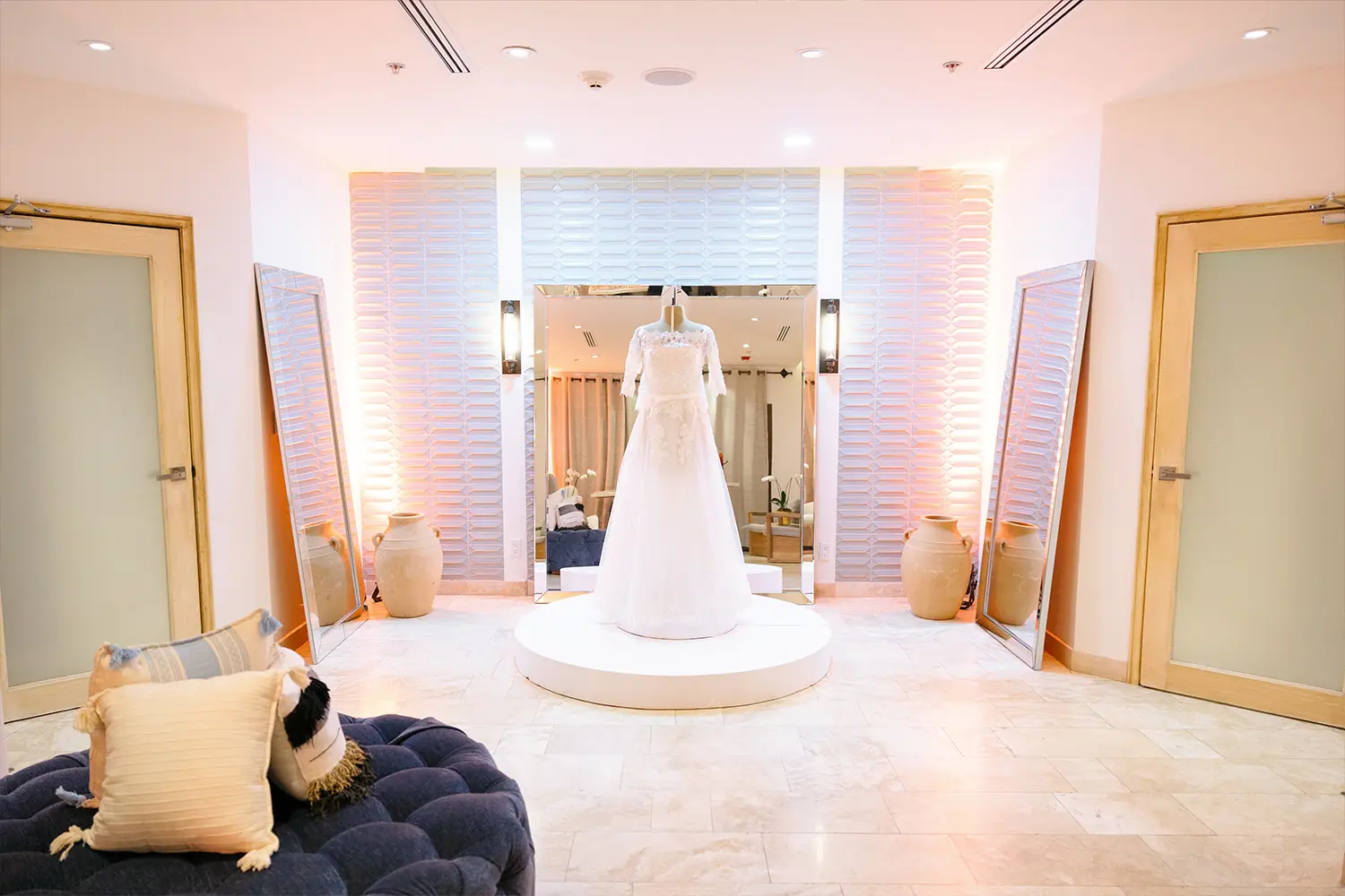 Un vestido de novia de encaje blanco exhibido en un pedestal en una suite nupcial suavemente iluminada con espejos y una decoración neutra.