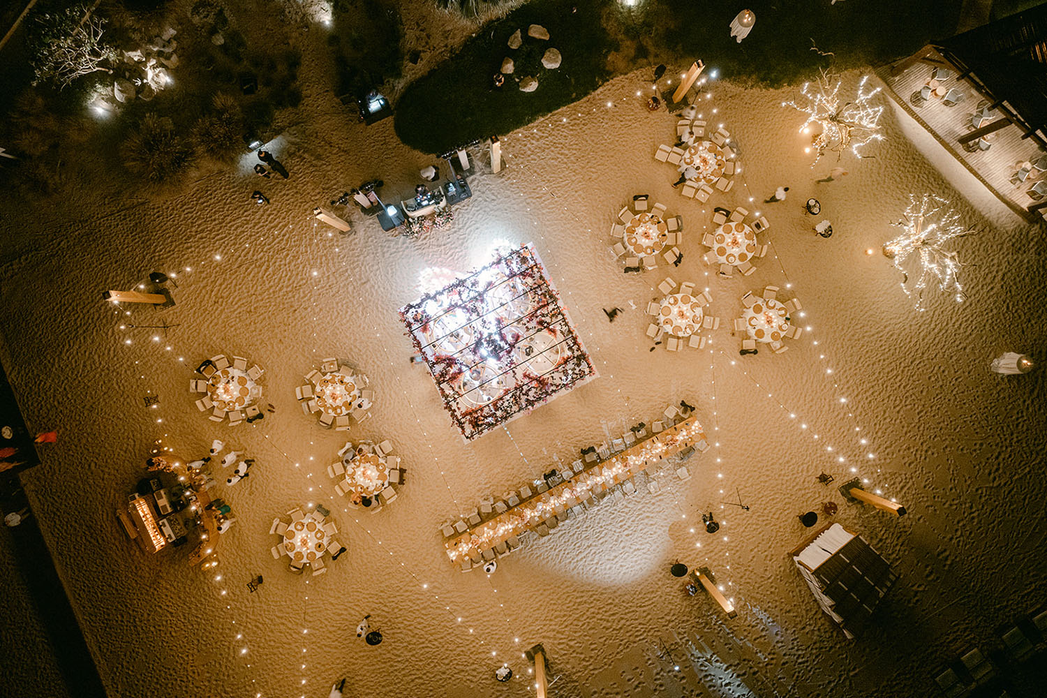Vista aérea de una elegante recepción de boda en la playa con mesas redondas, luces de cadena y una pista de baile central.