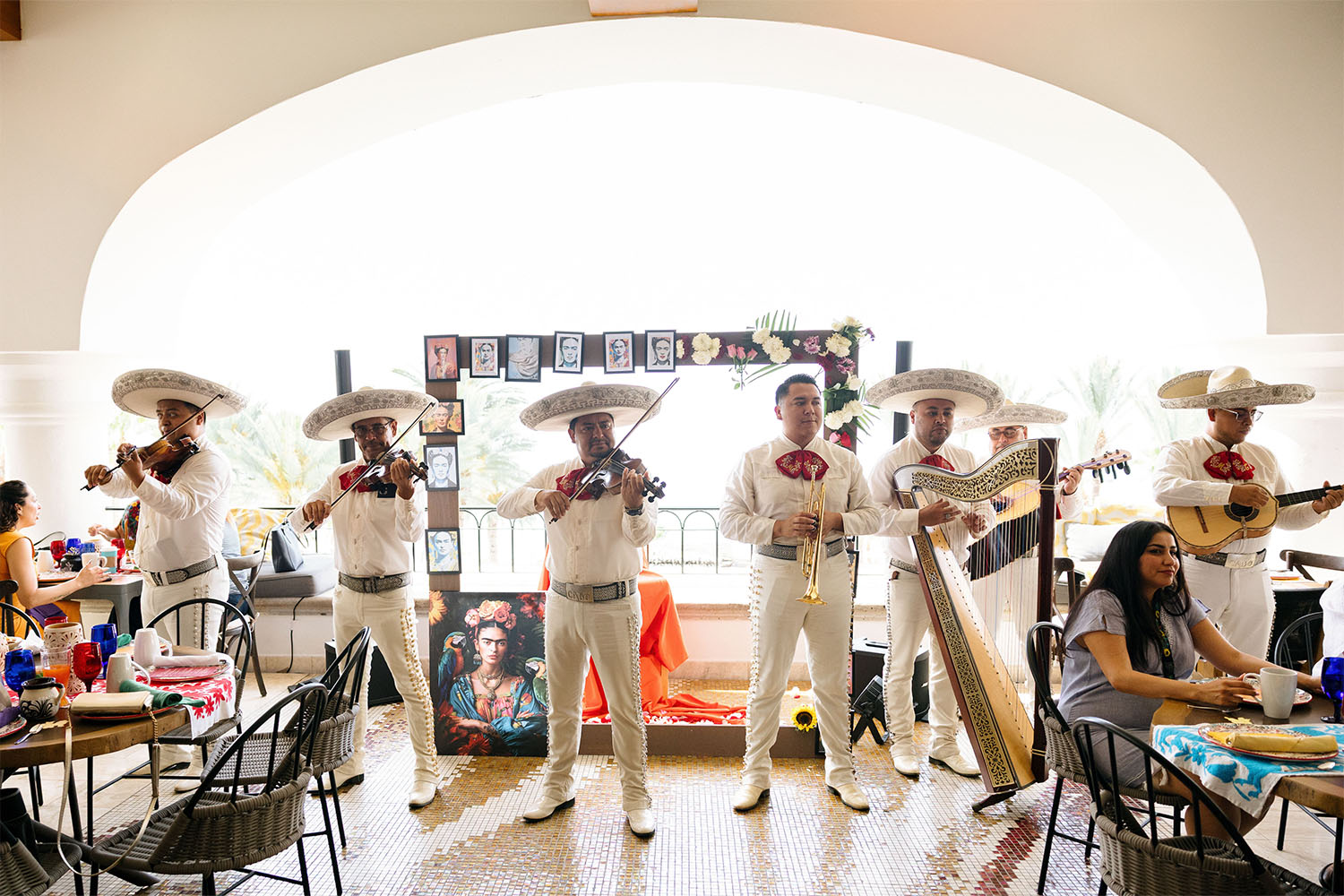 Banda de mariachis tocando en el interior con violines, trompeta, arpa y guitarras mientras los invitados cenan en mesas decoradas.