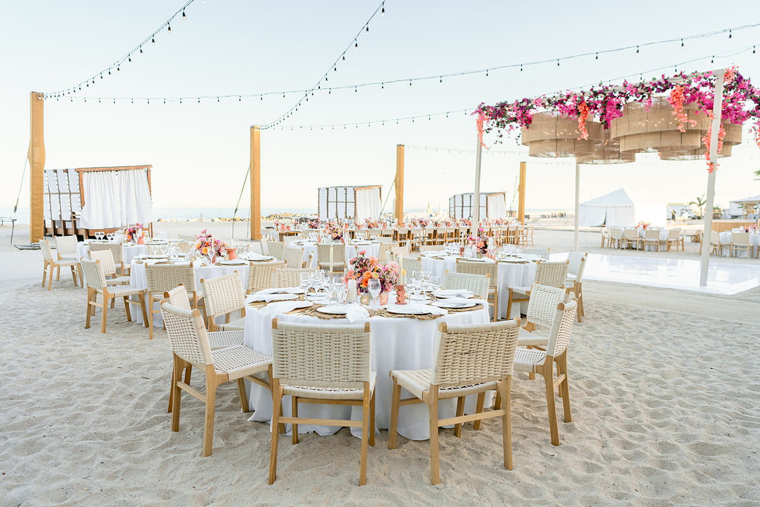 Recepción de boda frente a la playa con mesas redondas, mantelería blanca, centros de mesa florales y luces de cadena al atardecer.