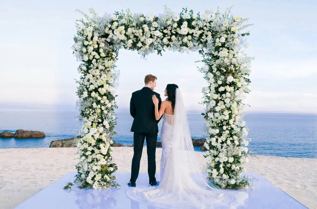 Bride and groom standing beneath a white floral arch during a beach wedding ceremony overlooking the ocean.