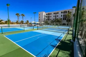 Canchas de pickleball al aire libre con superficies azules, cercas verdes y arquitectura de complejo enmarcada por palmeras.