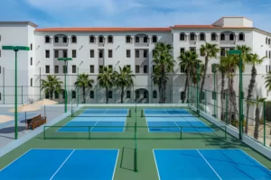 Outdoor pickleball courts with blue and green surfaces, palm trees, and Mediterranean-style resort building in the background.