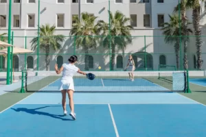 Dos mujeres jugando pickleball en una cancha al aire libre con palmeras y edificios de fondo.