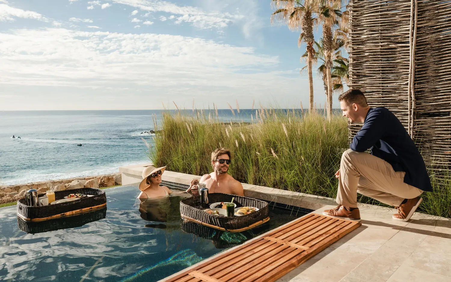 A couple enjoys a floating breakfast in a plunge pool overlooking the ocean while a Compadre Concierge team member checks in, at Hilton Los Cabos Beach & Golf Resort